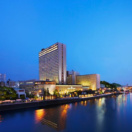 Rihga Royal Hotel Osaka, Vignette Collection By Ihg Exterior foto The photo shows a large hotel building situated near a body of water, likely a river. The scene is illuminated by soft evening lighting, giving the environment a tranquil, serene feel. In the background, there are various buildings and structures, su